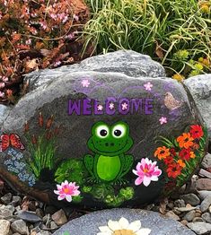 a rock with a frog painted on it sitting in front of some flowers and grass