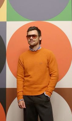 a man in an orange sweater and black pants standing against a wall with circles on it