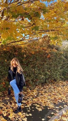 a woman standing in front of a tree with leaves on the ground next to her