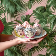 a woman holding three cupcakes with pink frosting on them in front of a palm tree