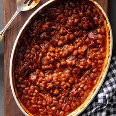 a large pot filled with baked beans on top of a wooden cutting board next to a knife and fork