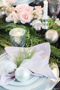 the table is set with white plates and silverware, pink roses, greenery and candlesticks