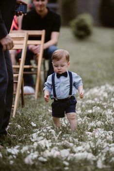 a little boy walking in the grass with a bow tie and suspenders around his neck