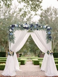 an outdoor wedding ceremony with white drapes and blue flowers on the arch, surrounded by hedges