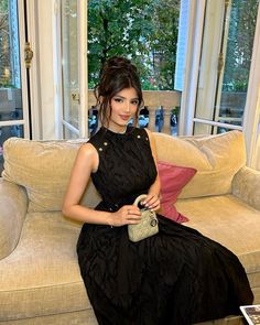 a woman in a black dress sitting on a couch holding a purse and posing for the camera