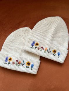 two white beanies with flowers on them sitting on top of a brown cloth covered surface