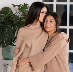 two women hugging each other in front of a window with potted plants behind them