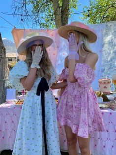 two women in dresses and hats standing next to each other near a table with food on it