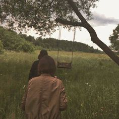 two people are walking through the grass in front of a swing chair that is suspended by a tree