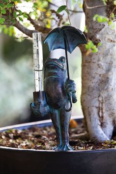 a statue of a frog holding an umbrella on top of a potted plant next to a tree