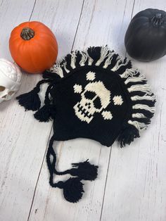 a skull and crossbones knitted hat sits on a white wooden surface next to two pumpkins