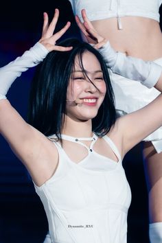 a woman with black hair wearing white clothes and holding her hands up in the air