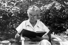 an old woman sitting on a chair reading a book in front of some bushes and trees