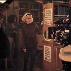 a woman standing in front of a book shelf with books on it and people behind her