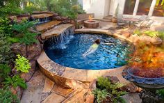 a man swimming in an outdoor pool surrounded by plants and rocks, next to a fire pit