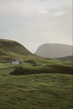 two white houses in the middle of a green field with mountains in the background and cloudy sky