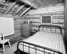 black and white photograph of an old log cabin bedroom with bed, dresser and chair