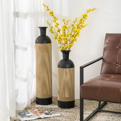 two black vases with yellow flowers in them next to a brown chair and window