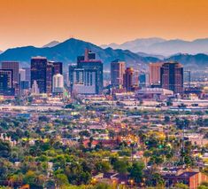 a city with mountains in the background and trees on the foreground, at sunset