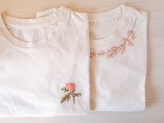 two white t - shirts with pink flowers on them sitting on a wooden table next to each other