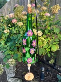 a green glass bottle with flowers painted on it hanging from a pole in front of a garden