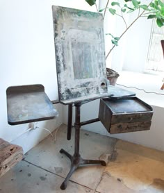 an old easel sits next to a table with two drawers on it and a potted plant in the corner