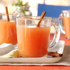 two mugs filled with orange juice and cinnamon sticks on a tablecloth covered tray