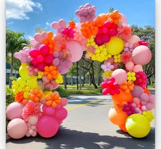 an arch made out of balloons and flowers on the side of a road with palm trees in the background