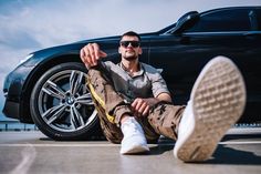 a man sitting on the ground in front of a black car with his feet up