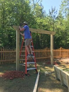 a man on a ladder working on a wooden structure in the yard with trees behind him