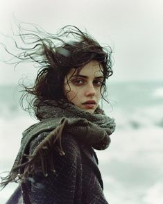 a woman standing on the beach with her hair blowing in the wind and looking at the camera