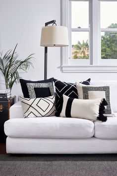 a white couch with black and white pillows in front of a large window next to a lamp