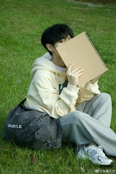 a person sitting in the grass with a notebook covering their face