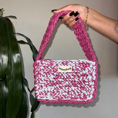 a hand holding a pink and white purse next to a plant with green leaves on it