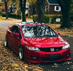 a red car parked on the side of a road in front of trees and leaves