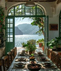 a dining table with plates and bowls on it in front of an open window overlooking the water