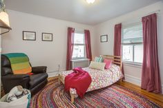 a living room with a bed, chair and rug in front of two windows on the wall