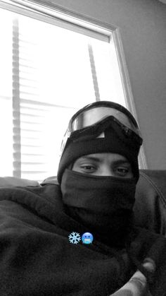 a person wearing a ski mask and goggles on their head, sitting in front of a window