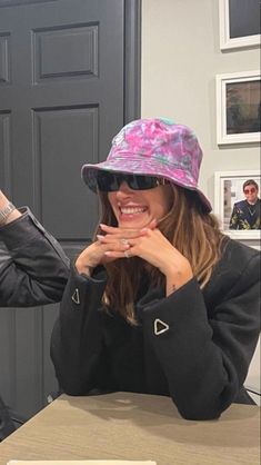 a woman sitting at a table wearing a pink hat and black jacket with her hands on her face