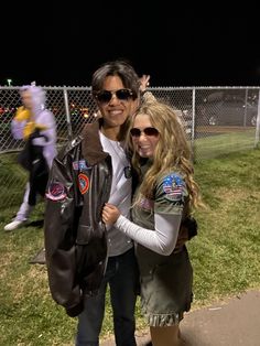 two people standing next to each other in front of a fence and field at night