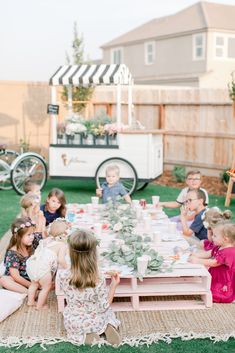 a group of children sitting around a table with food on it in the grass outside