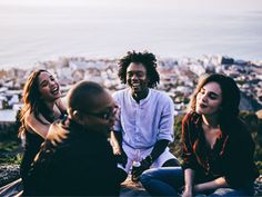 four people sitting on the ground laughing and looking at something in front of them with a cityscape in the background