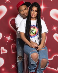 a man and woman sitting on top of a stool in front of a wall with hearts