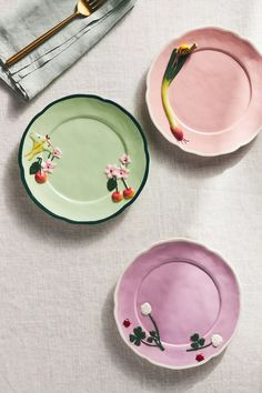 three plates with flowers painted on them sitting on a white table cloth next to a fork and knife