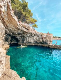 people are swimming in the blue water near some rocks and trees, while another person is standing at the end of the cave