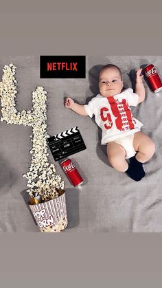 a baby laying on a bed next to a bucket of popcorn and a can of soda