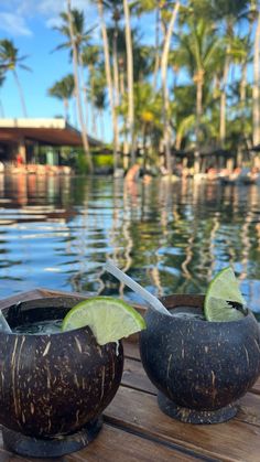 two coconuts with lime slices in them sitting on a wooden table next to the water