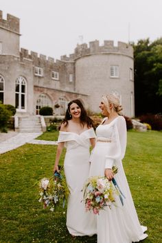 two women in white dresses standing next to each other on the grass near a castle