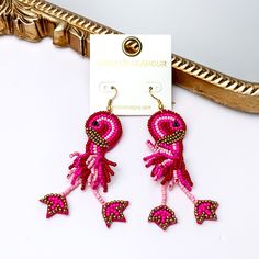 These earrings include multiple color pink beads and a gold detailing. Hanging from the bottom of the flamingo shaped beaded pendant has dangling legs. These earrings are pictured leaning agasint a gold mirror on a white background. Bohemian Jewelry For Summer Beach Party, Trendy Beaded Summer Jewelry, Trendy Beaded Earrings For The Beach, Bohemian Earrings For Summer Vacation, Trendy Beaded Drop Earrings For Vacation, Casual Summer Dangle Jewelry, Bohemian Pink Jewelry For Beach Party, Summer Vacation Drop Earrings, Trendy Earrings For The Beach Season