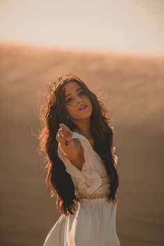 a woman standing in the desert wearing a white dress and holding her hand up to her face